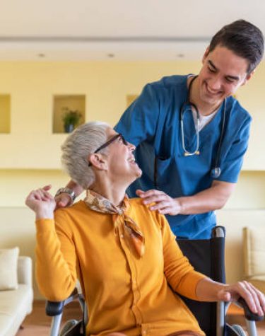 Senior Woman in Wheelchair is Having a Positive Communication with a Young Doctor in Blue Uniform who is Giving her Support and Pushing her Through the House.